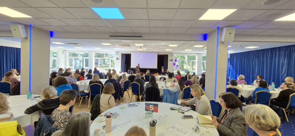 A wide-angle view of a large workshop space with attendees seated at numerous round white-clothed tables. A presenter stands at the far end of the room in front of a projector screen. The room is brightly lit by large windows and square ceiling lights, with blue vertical lighting accents on the structural pillars.