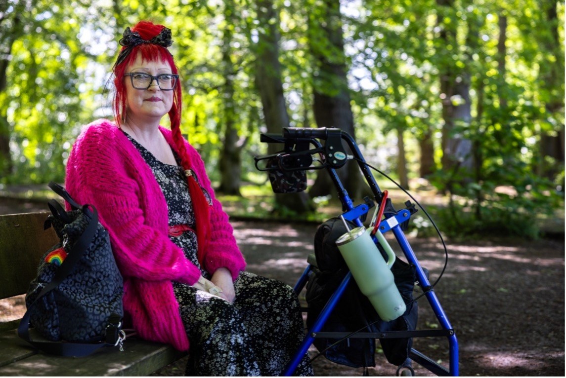 Photograph from the Claiming the Normal project: a disabled model sits on a wooden bench in a leafy park, wearing glasses, a bright pink cardigan over a dark patterned dress, with a blue rollator beside him.