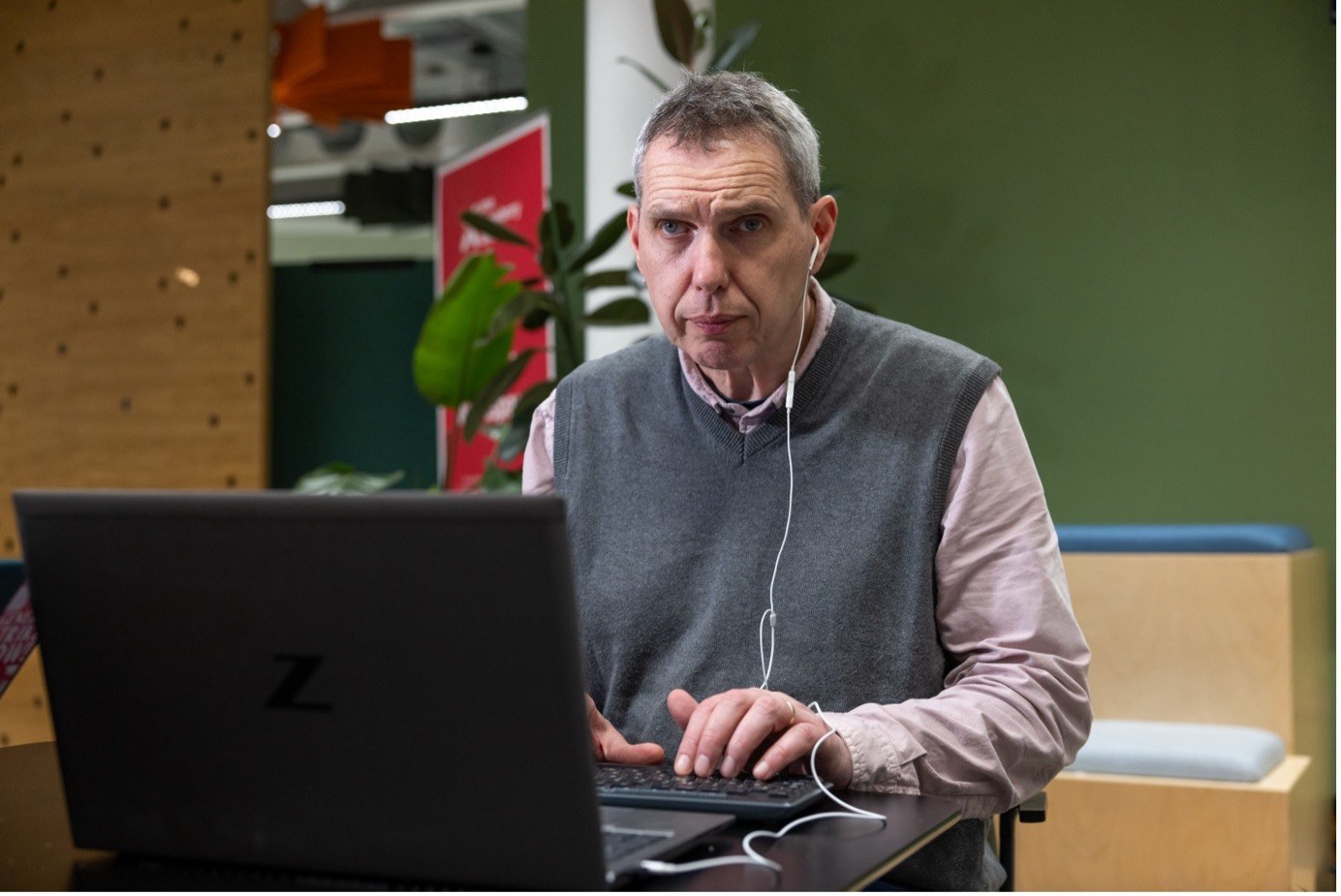 A man with short grey hair, wearing earphones and a grey vest over a pink shirt, sits at a desk working on a laptop in an office setting.