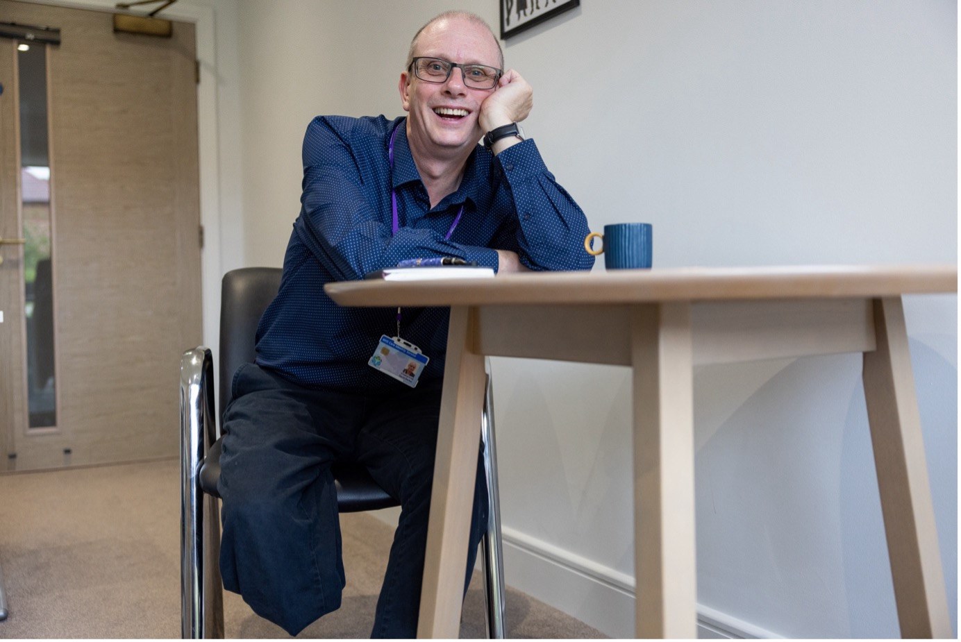 Photograph from the Claiming the Normal project: a bald man with glasses, wearing a dark blue shirt and a lanyard, sits at a table with a blue mug, smiling and resting his head on one hand.